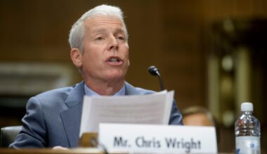 Chris Wright sitzt bei einer Veranstaltung hinter einem Mikrofon. Vor ihm steht sein Namensschild.