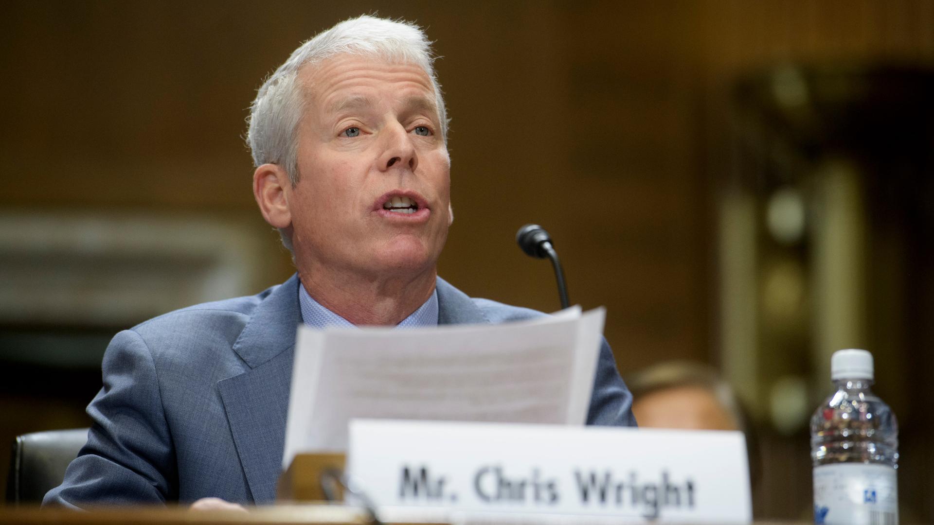 Chris Wright sitzt bei einer Veranstaltung hinter einem Mikrofon. Vor ihm steht sein Namensschild.