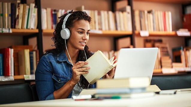Junge Frau mit Kopfhören sitzt in einer Bibliothek vor einem Laptop und blättert in einem Buch.