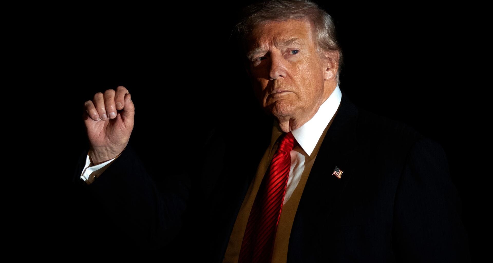 Donald Trump im Anzug und mit roter Krawatte wird in Dunkelheit vom Blitzlicht einer Kamera beleuchtet, während er mit der rechten Hand gestikuliert