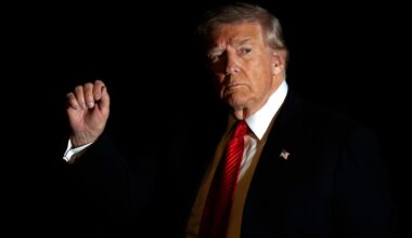 Donald Trump im Anzug und mit roter Krawatte wird in Dunkelheit vom Blitzlicht einer Kamera beleuchtet, während er mit der rechten Hand gestikuliert