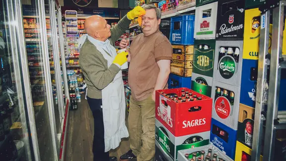 Zwei Männer in einerm Kiosk. Der eine säubert den anderen mit einem Tuch.