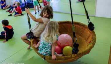 Zwei Kinder spielen Indoor auf einer Schaukel, dahinter sitzen andere Kinder auf dem Boden