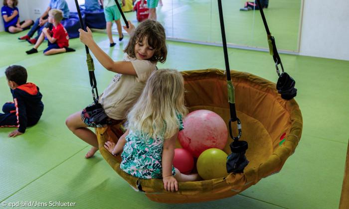 Zwei Kinder spielen Indoor auf einer Schaukel, dahinter sitzen andere Kinder auf dem Boden