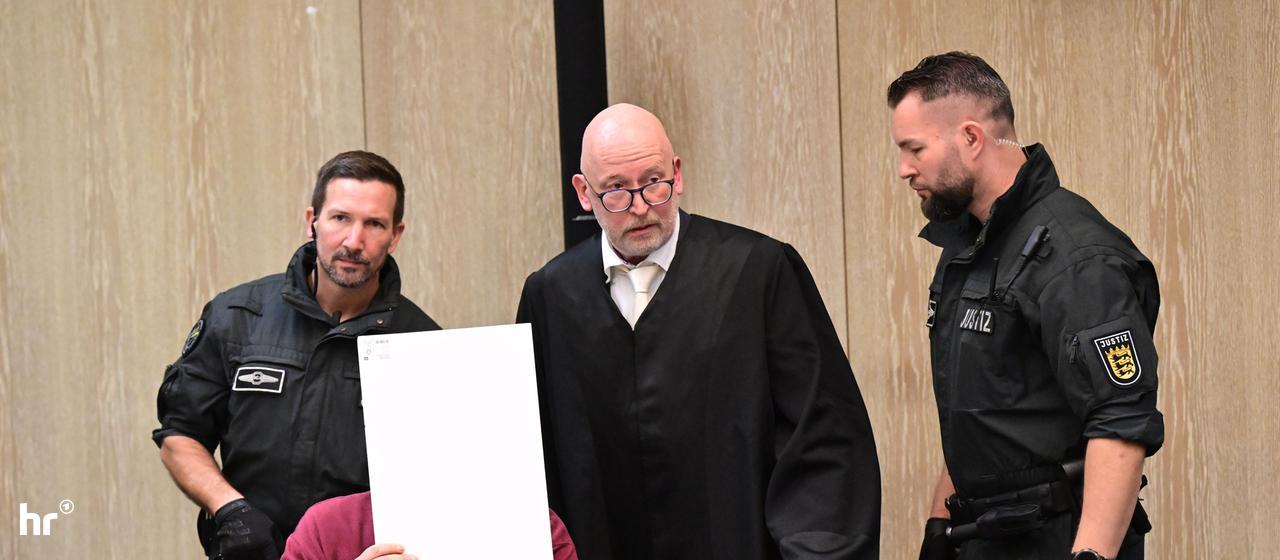 Der Angeklagte in einem Mordprozess nach einer Todesfahrt von Mannheim sitzt mit einem Aktendeckel vor seinem Gesicht im Gerichtssaal, rechts neben ihm steht sein Verteidiger.