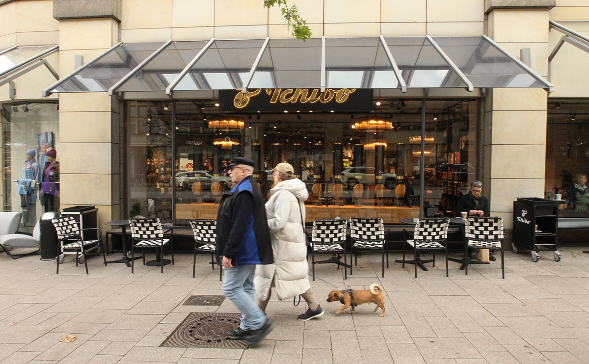 Tchibo-Filiale bei Tageslicht, zwei Spaziergänger laufen daran vorbei.