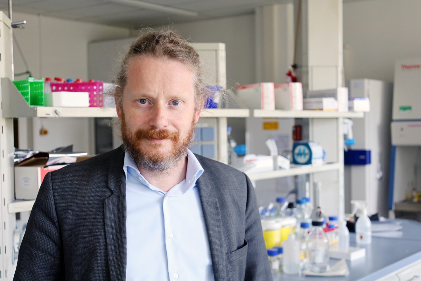 Ein Mann mit Bart trägt einen grauen Anzug und steht in einem Labor mit Regalen und Geräten.