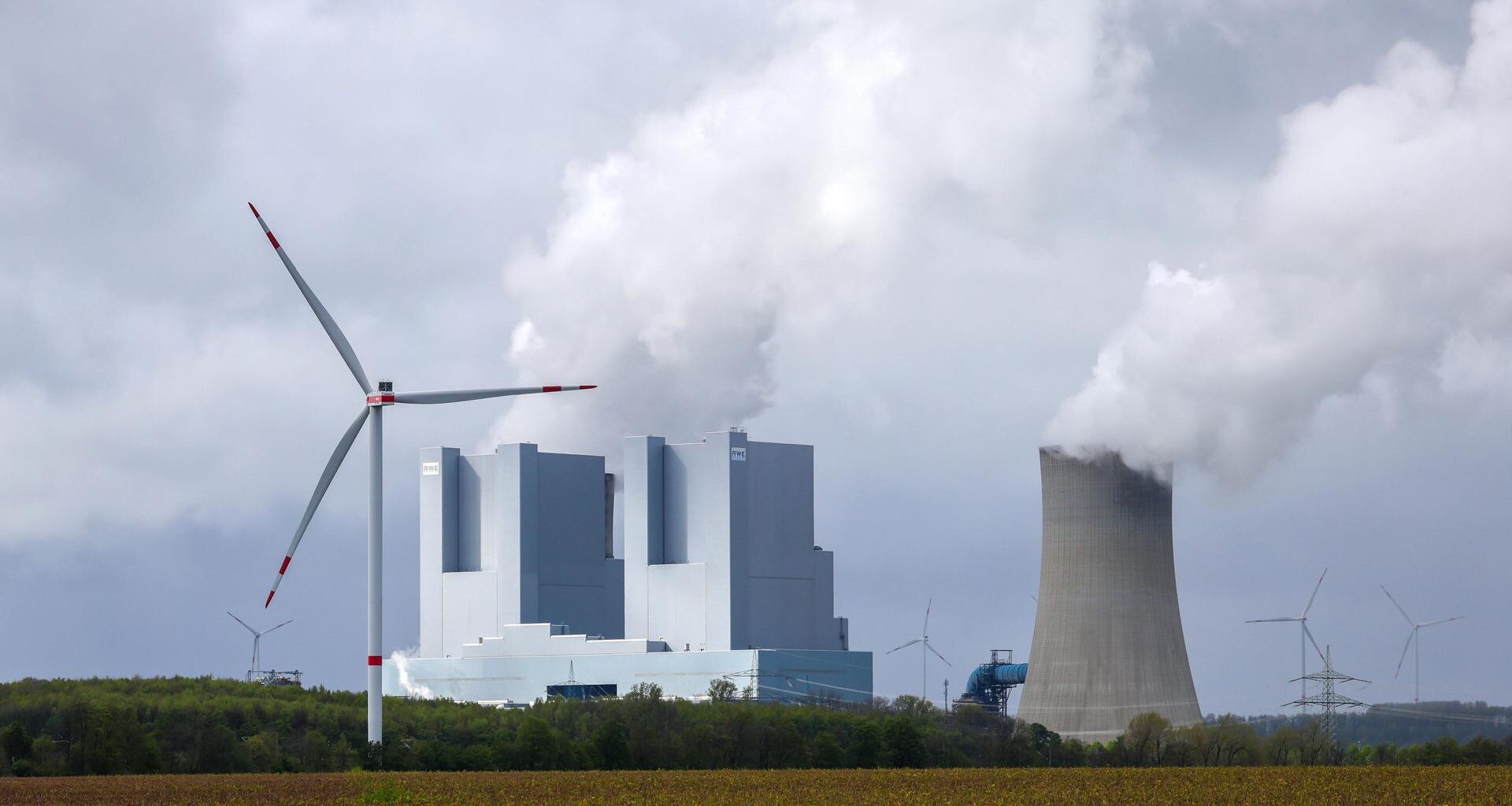 Hinter einem Feld steht das RWE-Kraftwerk in Grevenbroich mit Kühltürmen aus denen Dampf in den Himmel steigt. Davor und dahinter stehen Windräder.