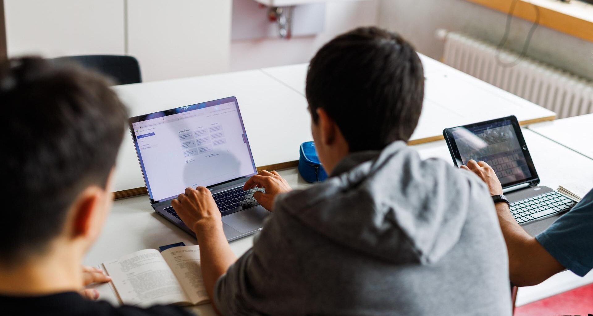 Drei Schüler von hinten an einer Schulbank; zwei von ihnen schauen in ein Tablet.