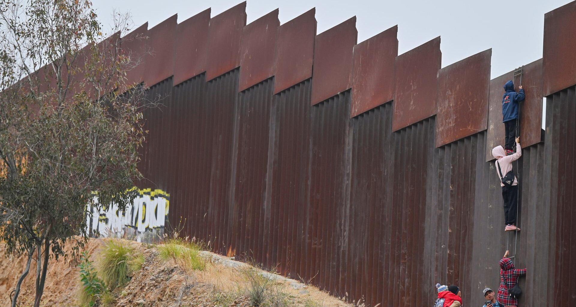 Eine kleine Gruppe von Migranten, darunter mindestens ein Kind, versucht, mit einer Leiter über die Grenzmauer in die USA zu gelangen.