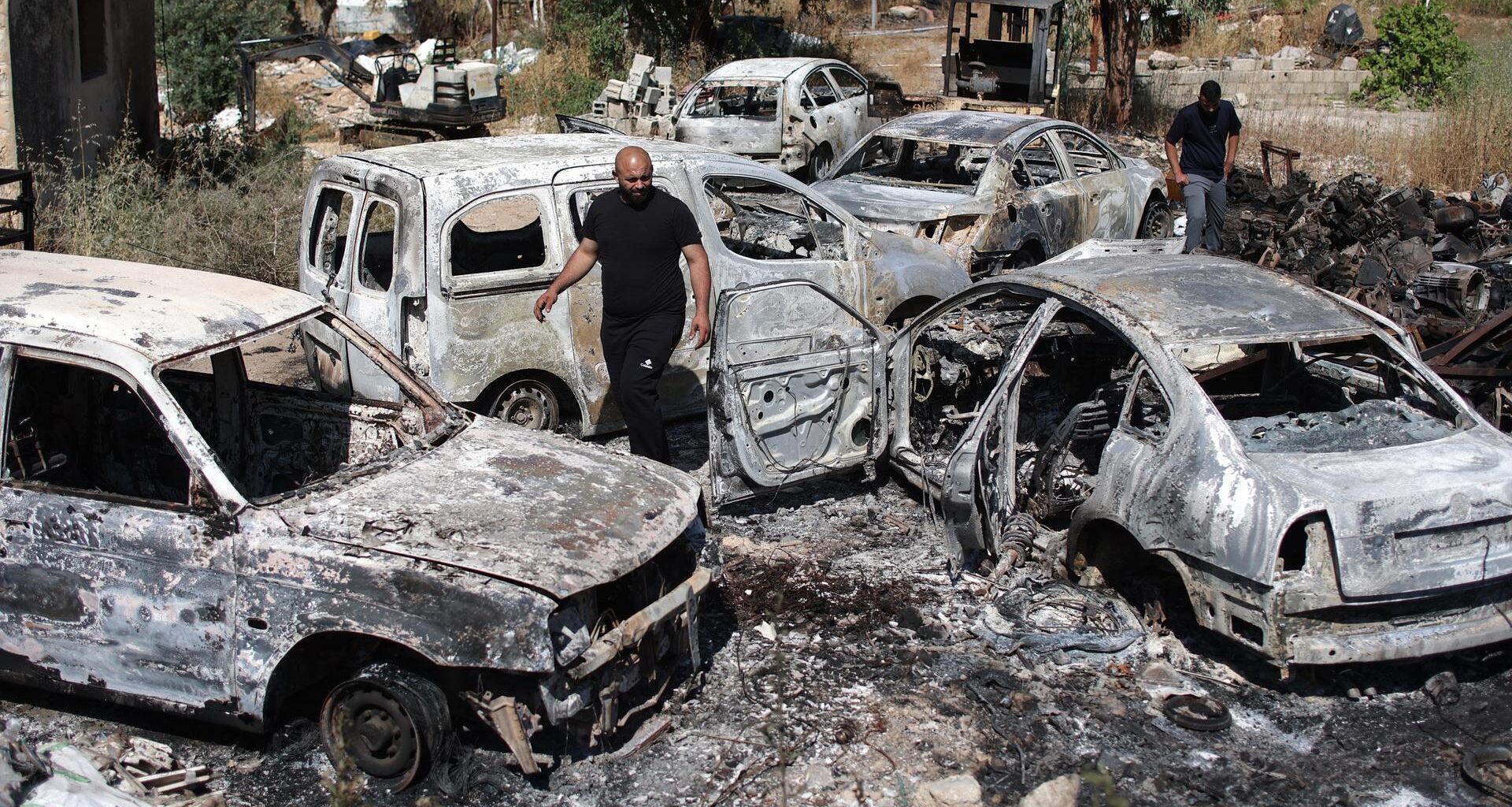 Zwei Männer laufen zwischen fünf völlig ausgebrannten Fahrzeugen auf einem Gelände nahe einer Straße herum.
