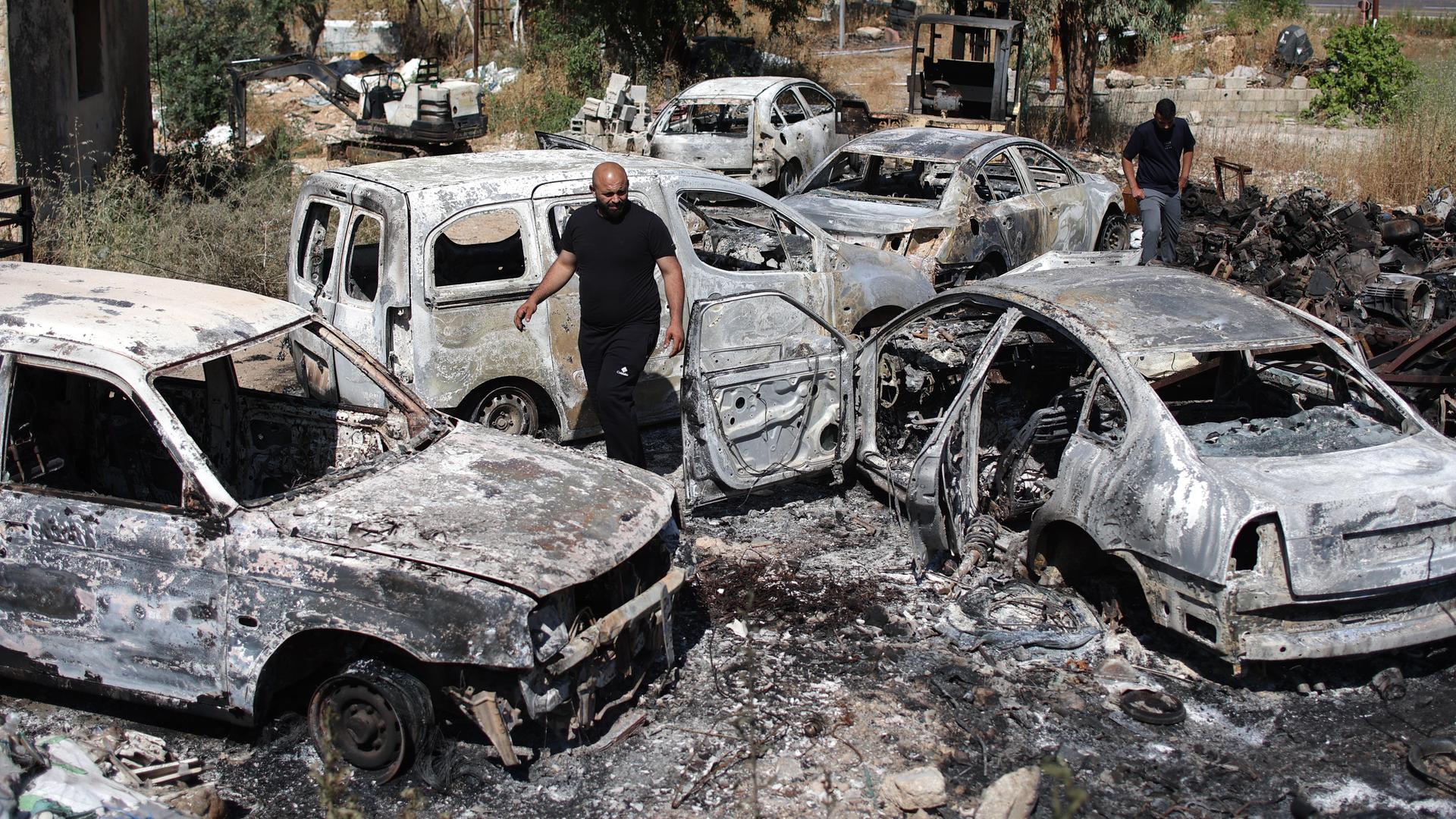 Zwei Männer laufen zwischen fünf völlig ausgebrannten Fahrzeugen auf einem Gelände nahe einer Straße herum. 