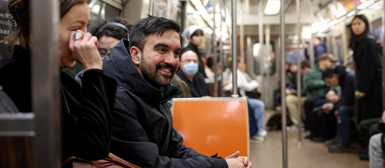 Zohran Mamdani sitzt lächelnd in der U-Bahn. | REUTERS Zohran Mamdani sitzt lächelnd in der U-Bahn.