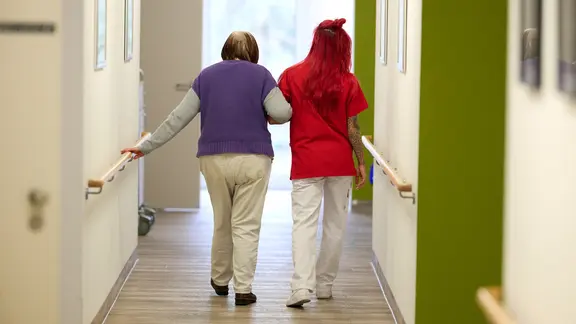 Eine Pflegefachkraft geht Arm in Arm mit einer Patientin über einen Flur. (Themenbild) 