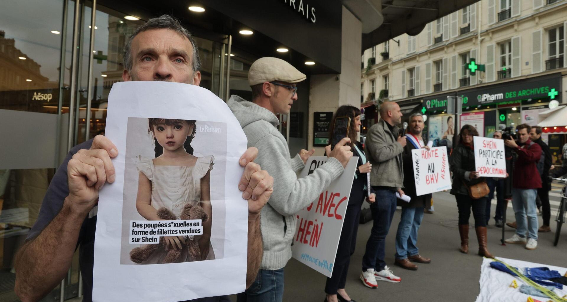 Mehrere Menschen stehen vor dem Eingang eines kaufhauses und halten beschriftete Protestschilder sowie vergrößerte Fotos der Sexpuppen.