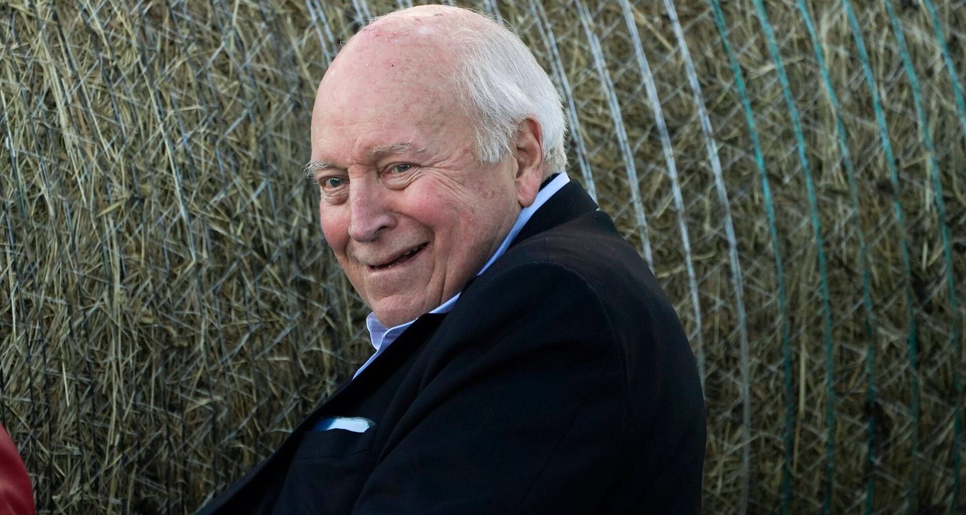 Richard Cheney sitzt seitlich, schaut in die Kamera und lächelt.