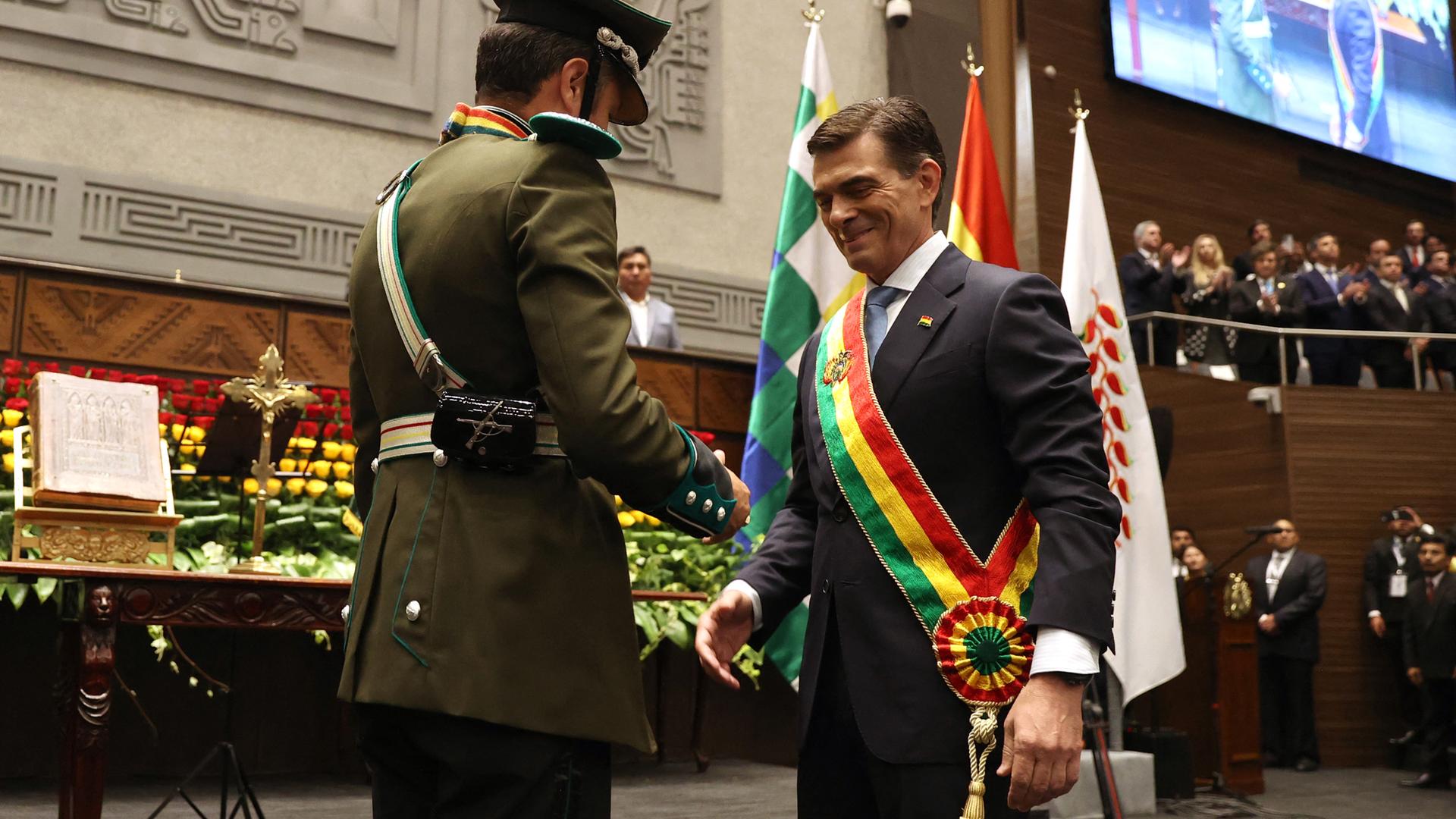 Der lächelnde Paz trägt die Schärpe, die er von einem Armeeangehörigen in Paradeuniform überreicht bekommen hat. Dahinter eine vollbesetzte Tribüne.