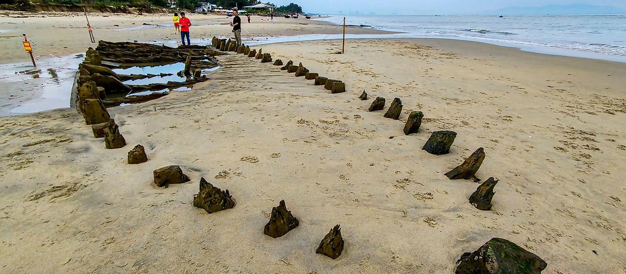 Personen untersuchen das hölzerne Wrack eines Schiffes auf einem Strand.