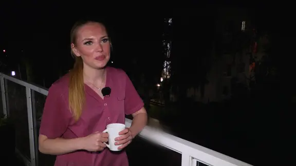 Eine junge Frau mit blonden Haaren und in einem brombeerfarbenen Kittel hält eine Tasse in den Händen.