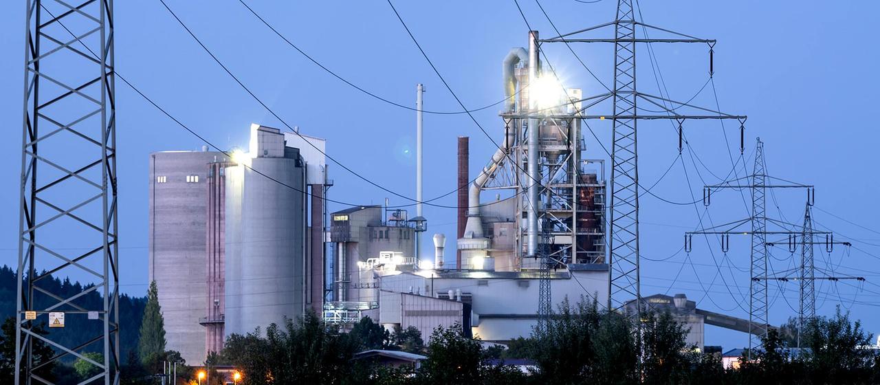 Strommasten stehen im Morgenlicht vor einem Zementwerk. | picture alliance/dpa Strommasten stehen im Morgenlicht vor einem Zementwerk.