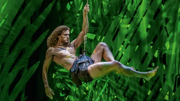 Szenenbild aus dem Musical Tarzan: Ein Mann mit langen Haaren und Lendenschurz schwingt ein einem Seil, im Hintergrund Grün.