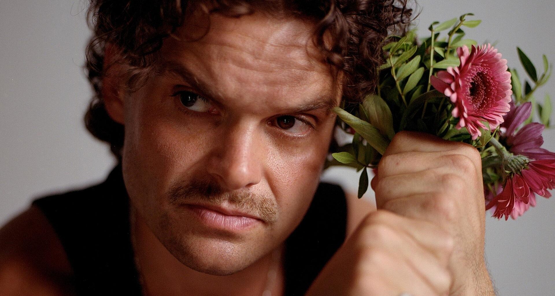 Ein Mann mit braunen Locken und Schnauzbart schaut ernst aus dem Bild hinaus. Neben dem Kopf hält er in seinen Fäusten einen Blumenstrauß mit rosa Gerbera. Er trägt ein schwarzes Tanktop und silbernen Schmuck.