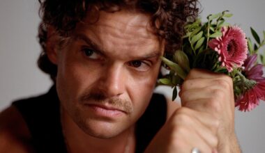 Ein Mann mit braunen Locken und Schnauzbart schaut ernst aus dem Bild hinaus. Neben dem Kopf hält er in seinen Fäusten einen Blumenstrauß mit rosa Gerbera. Er trägt ein schwarzes Tanktop und silbernen Schmuck.