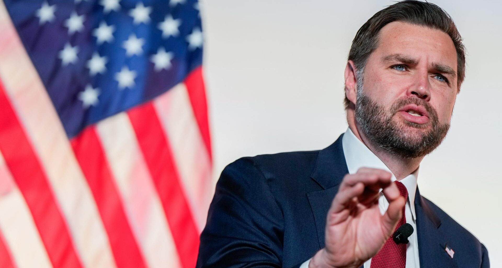 Der bärtige Vance spricht vor einer amerikanischen Flagge. Im Hintergrund eine weiße Wand. Er trägt ein schwarzes Jackett.
