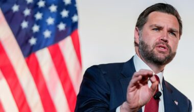 Der bärtige Vance spricht vor einer amerikanischen Flagge. Im Hintergrund eine weiße Wand. Er trägt ein schwarzes Jackett.