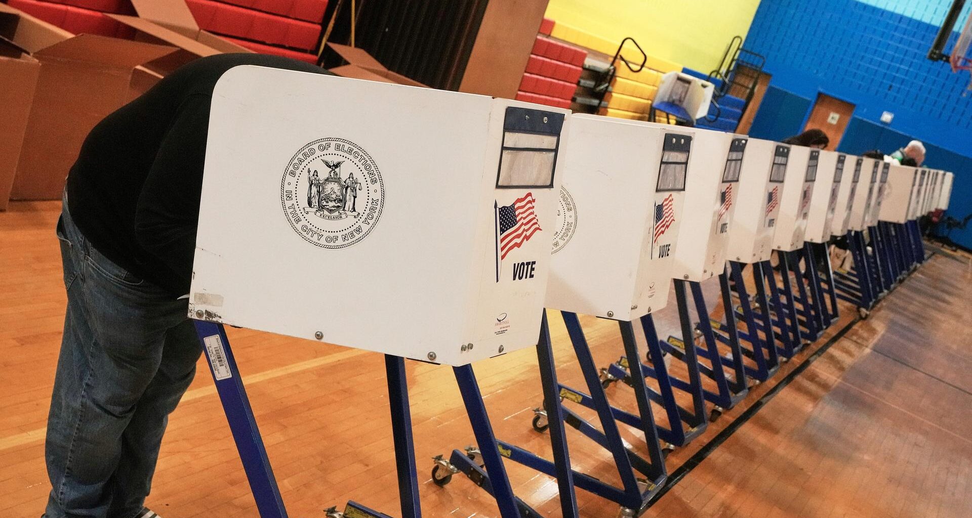 In einer langen Reihe von Wahlkabinen stehen drei Menschen, um ihre Stimme abzugeben.