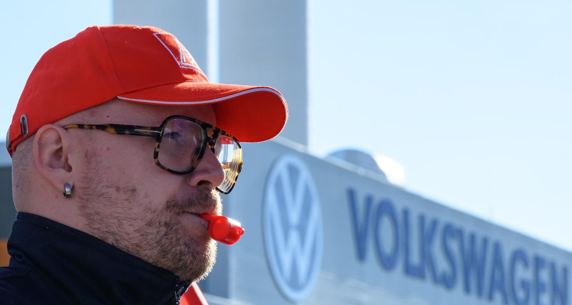 Ein Beschäftigter von Volkswagen hat eine rote Trillerpfeife im Mund und eine rote Baseball-Cappy auf dem Kopf und steht vor dem Volkswagen-Schriftzug