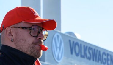 Ein Beschäftigter von Volkswagen hat eine rote Trillerpfeife im Mund und eine rote Baseball-Cappy auf dem Kopf und steht vor dem Volkswagen-Schriftzug