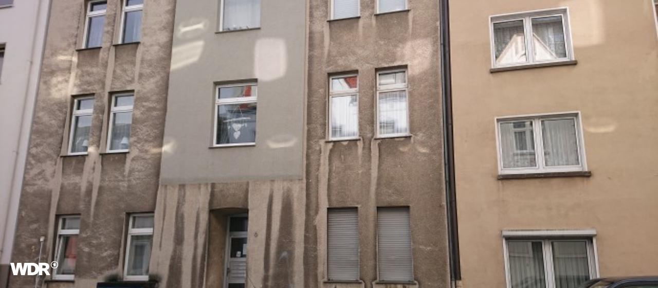 Ein graues Mehrfamilienhaus mit geparkten Autos vor der Tür