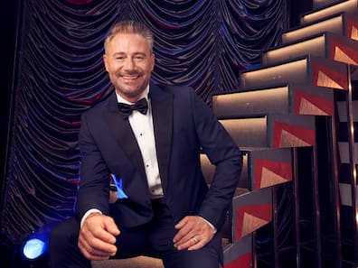 Sasha in elegantem Showanzug lachend auf einer Treppe sitzend, Porträt für das Große Adventskonzert Dresden 2025.