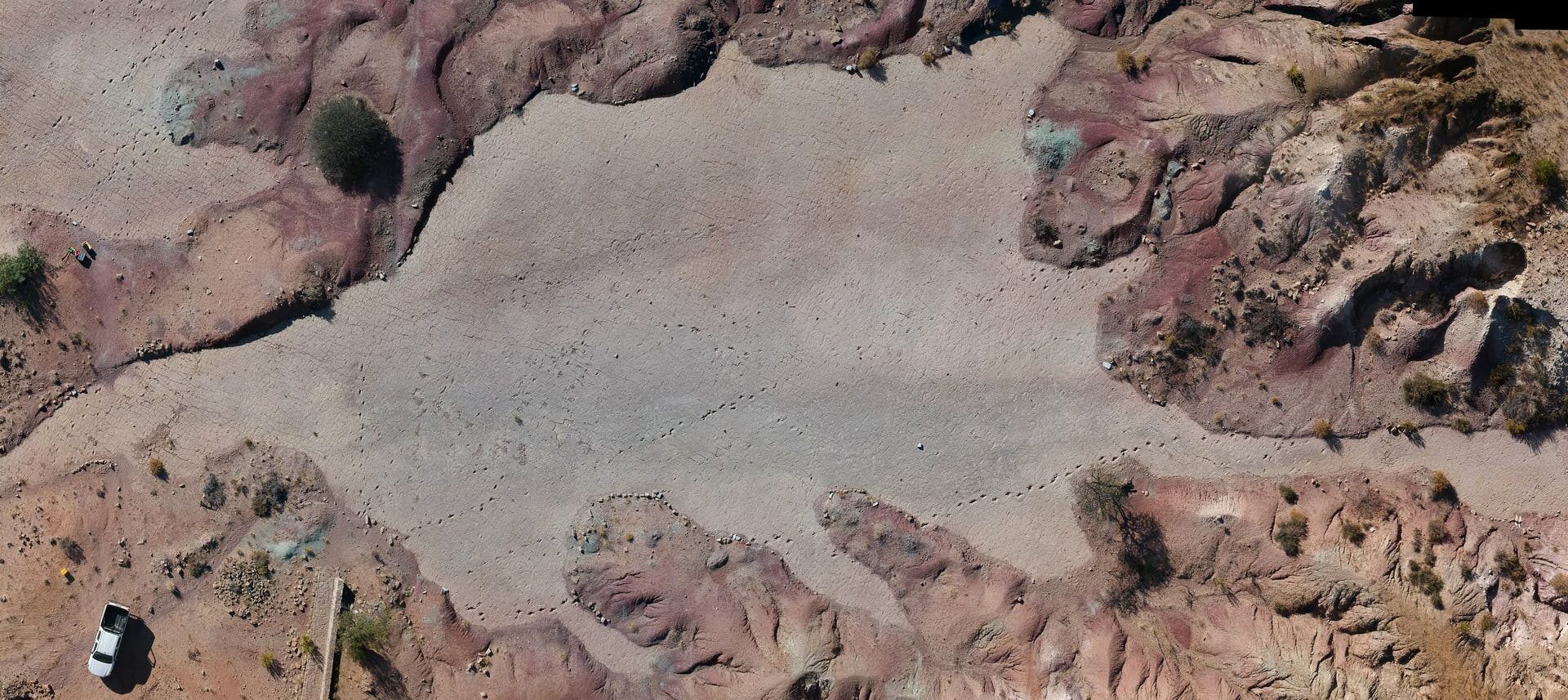 Luftaufnahme einer trockenen, felsigen Landschaft mit rötlichen und bräunlichen Tönen. In der unteren linken Ecke ist ein weißes Fahrzeug zu sehen. Die Oberfläche zeigt Spuren von Theropoden.