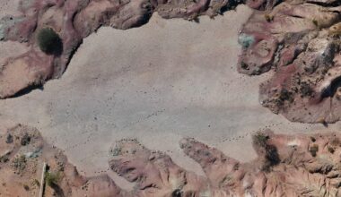 Luftaufnahme einer trockenen, felsigen Landschaft mit rötlichen und bräunlichen Tönen. In der unteren linken Ecke ist ein weißes Fahrzeug zu sehen. Die Oberfläche zeigt Spuren von Theropoden.