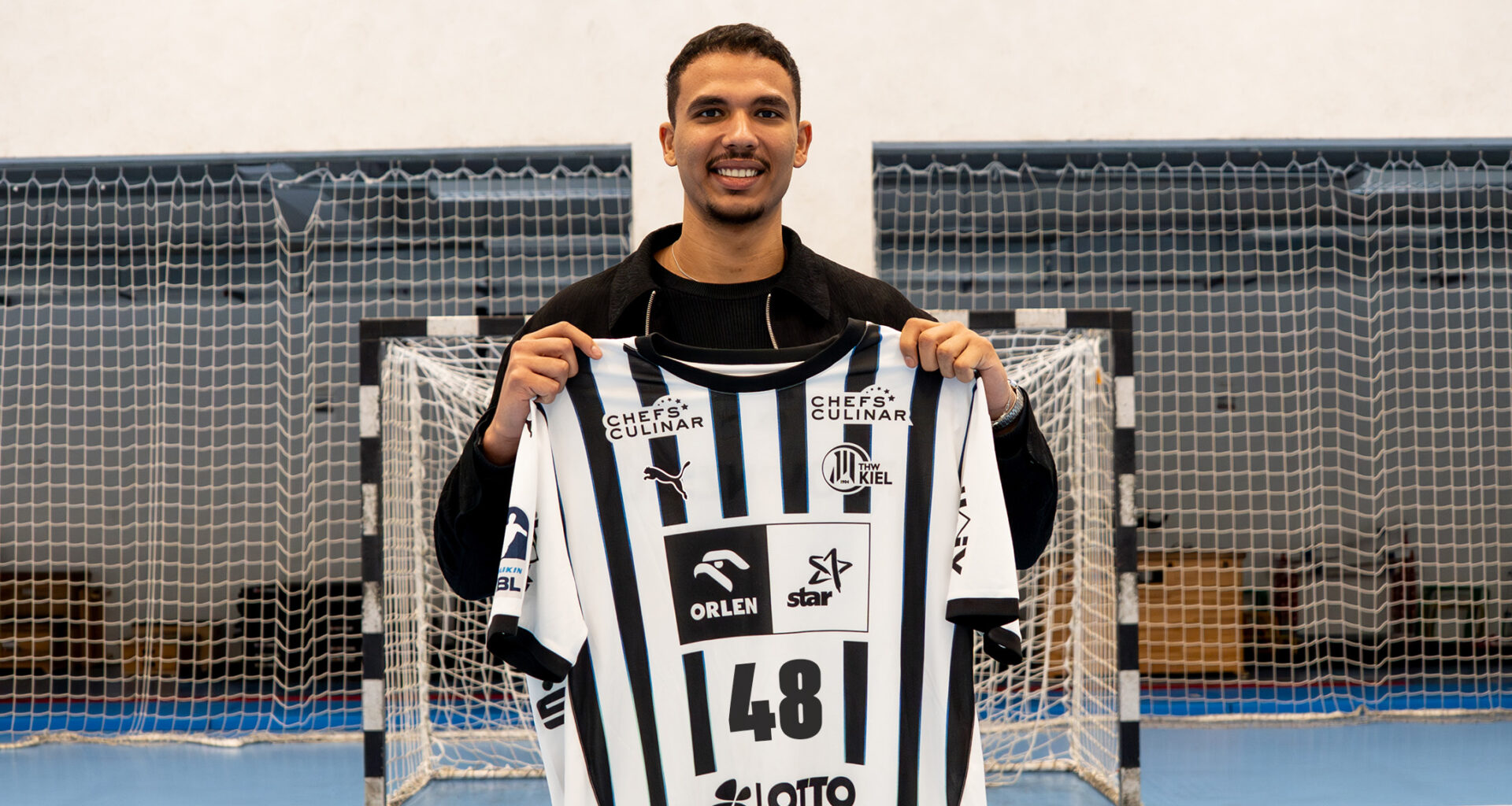 Ein lächelnder Mann steht drinnen auf einem Sportplatz und hält ein schwarz-weiß gestreiftes Trikot mit der Nummer 48 vor ein Tornetz.