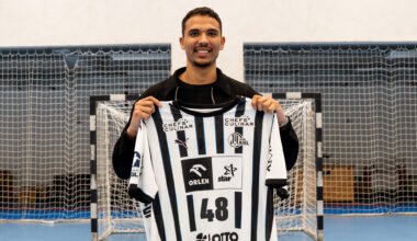 Ein lächelnder Mann steht drinnen auf einem Sportplatz und hält ein schwarz-weiß gestreiftes Trikot mit der Nummer 48 vor ein Tornetz.