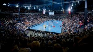 Eine große Halle voller Zuschauer, die ein Handballspiel verfolgen. Helle Lichter beleuchten das blaue Spielfeld, die Spieler sind in Aktion und die Schiedsrichter in der Nähe. Die Tribünen sind voll besetzt, und es herrscht eine energiegeladene und lebhafte Atmosphäre.