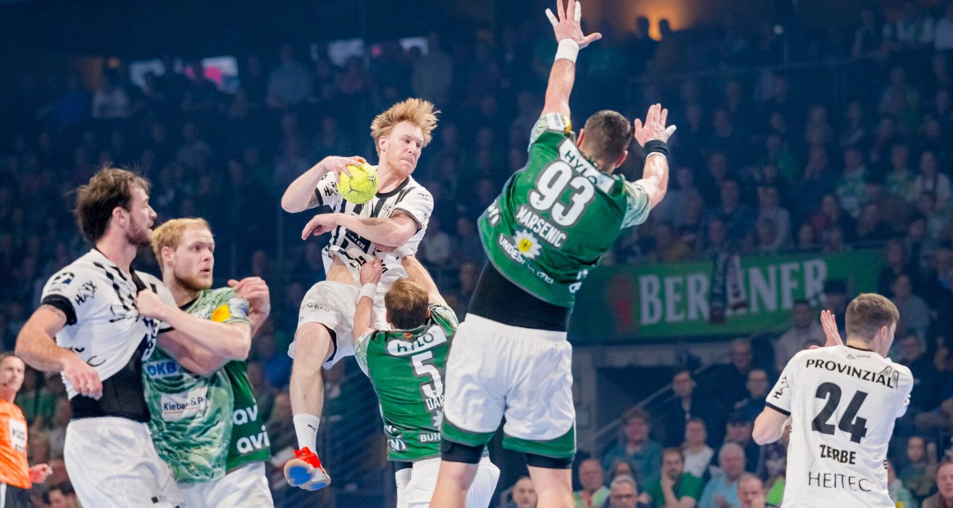 Ein Handballspieler in einem weißen Trikot springt in der Luft, um den Ball zu werfen, während er von gegnerischen Spielern in grünen Trikots umringt wird, die versuchen, ihn während eines intensiven Spiels zu blockieren.