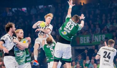 Ein Handballspieler in einem weißen Trikot springt in der Luft, um den Ball zu werfen, während er von gegnerischen Spielern in grünen Trikots umringt wird, die versuchen, ihn während eines intensiven Spiels zu blockieren.