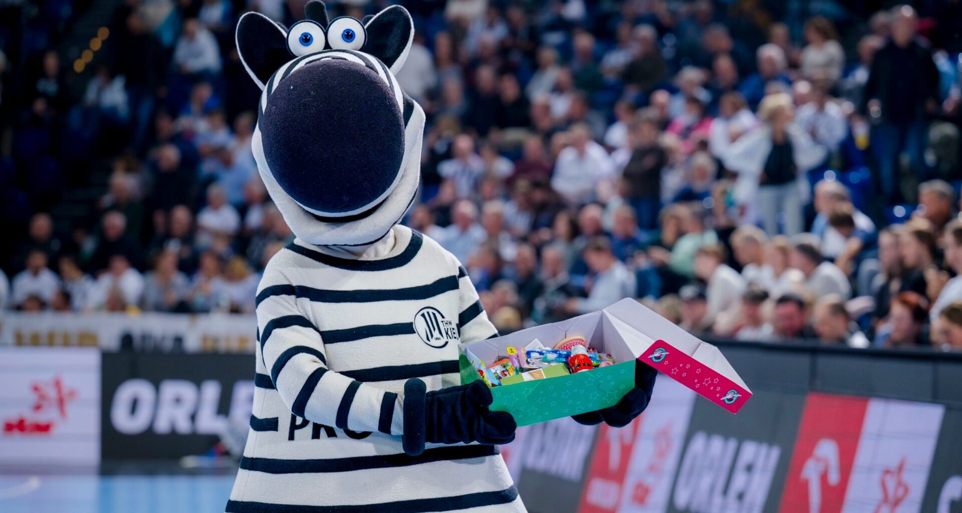 Eine Person in einem Zebra-Maskottchen-Kostüm hält eine offene Schachtel mit bunten Gegenständen in der Hand und steht auf einem Sportplatz mit einer unscharfen Menschenmenge im Hintergrund.