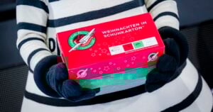 Eine Person mit schwarzen Handschuhen und einem gestreiften Outfit hält einen rot-grünen Schuhkarton mit der Aufschrift WEIHNACHTEN IM SCHUHKARTON und dem Logo von Samaritan's Purse Operation Christmas Child.