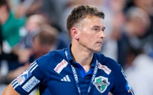 Ein Mann, der ein blaues Sporthemd mit Sponsorenlogos und ein Schlüsselband trägt, steht in einer Halle und schaut mit ernster Miene zur Seite. Der unscharfe Hintergrund suggeriert Aktivität, möglicherweise im Zusammenhang mit dem THW Kiel Saison 2025/2026 Handball-Bundesliga Vorbericht.