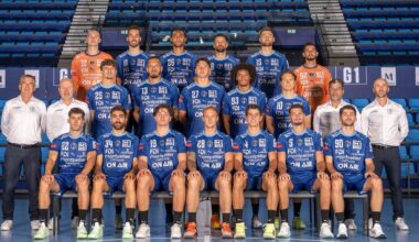 Eine Herren-Handballmannschaft stellt sich für ein Gruppenfoto in einem Stadion auf. Die Spieler in blauen Trikots sitzen und stehen, flankiert von Trainern in weißen Hemden und leeren blauen Sitzen im Hintergrund.
