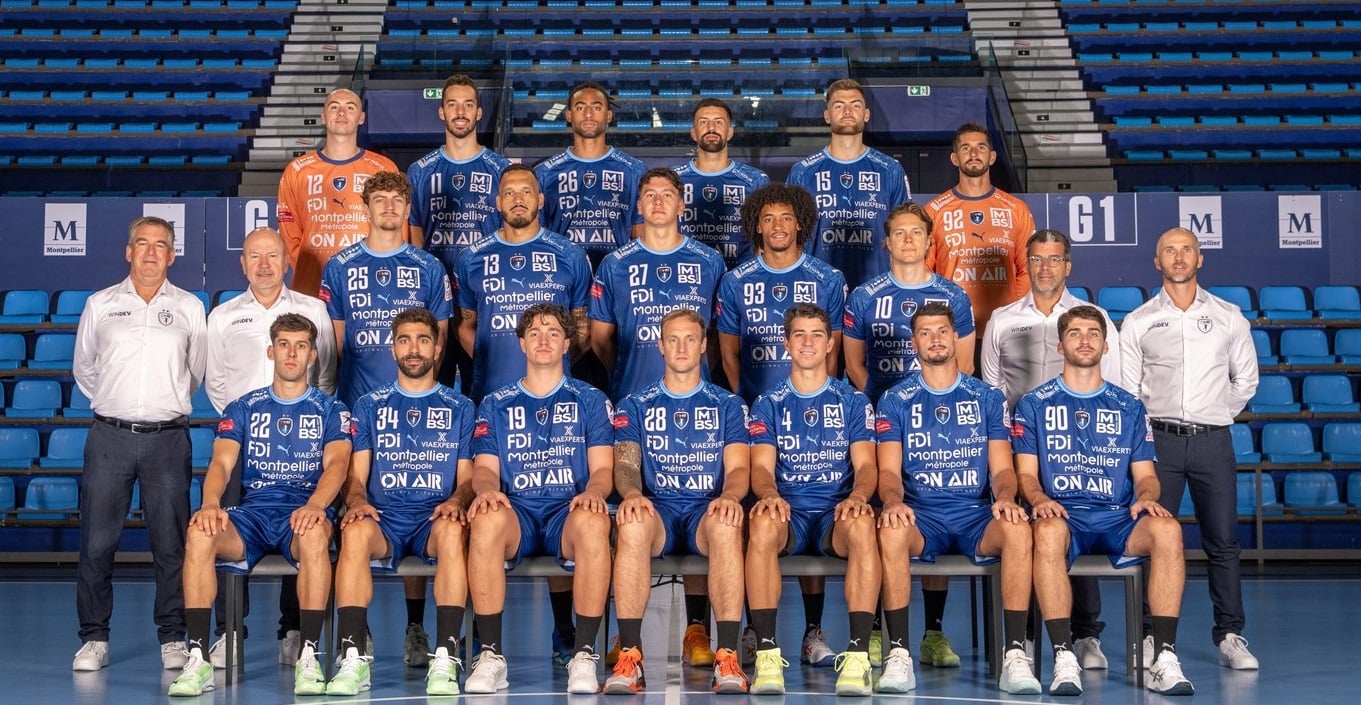 Eine Herren-Handballmannschaft stellt sich für ein Gruppenfoto in einem Stadion auf. Die Spieler in blauen Trikots sitzen und stehen, flankiert von Trainern in weißen Hemden und leeren blauen Sitzen im Hintergrund.