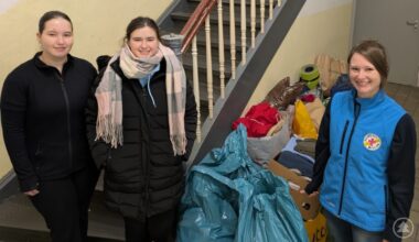Drei junge Frauen stehen neben mehreren großen Taschen und Kartons mit gespendeter Kleidung im Treppenhaus. Zwei tragen schwarze Winterjacken, eine (rechts) trägt eine blaue Weste mit Logo der Bahnhofsmission. Im Hintergrund sieht man eine Treppe.