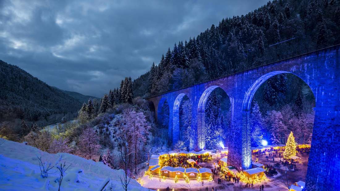 Verschneiter Weihnachtsmarkt unter einem blau beleuchteten Eisenbahn-Viadukt.