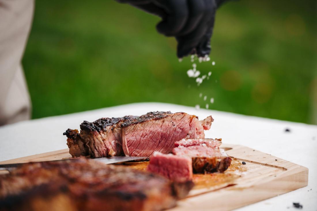 Chef salting juicy grilled steak on wooden cutting board. 
