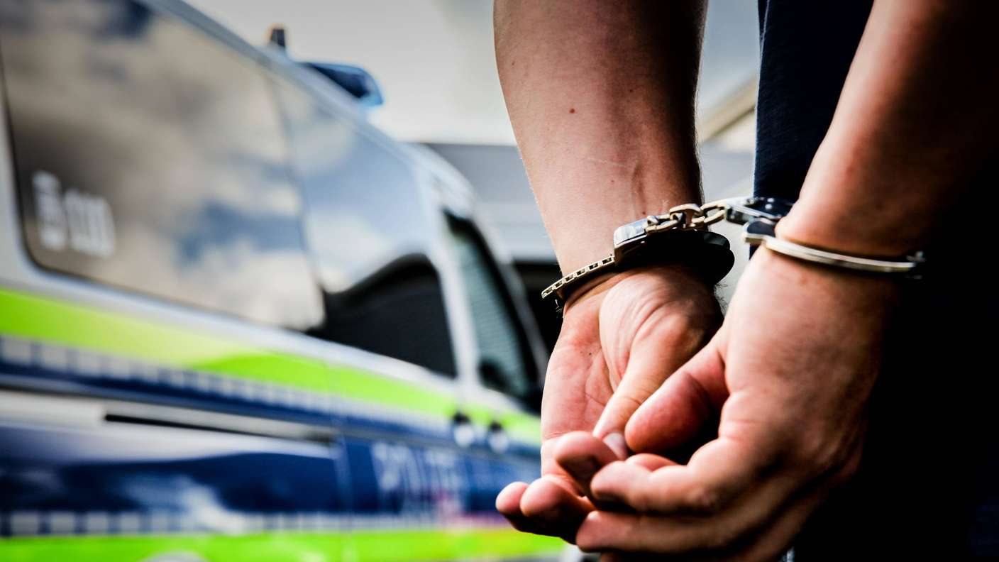 Ein Mann mit Handschellen vor einem Polizeifahrzeug. 